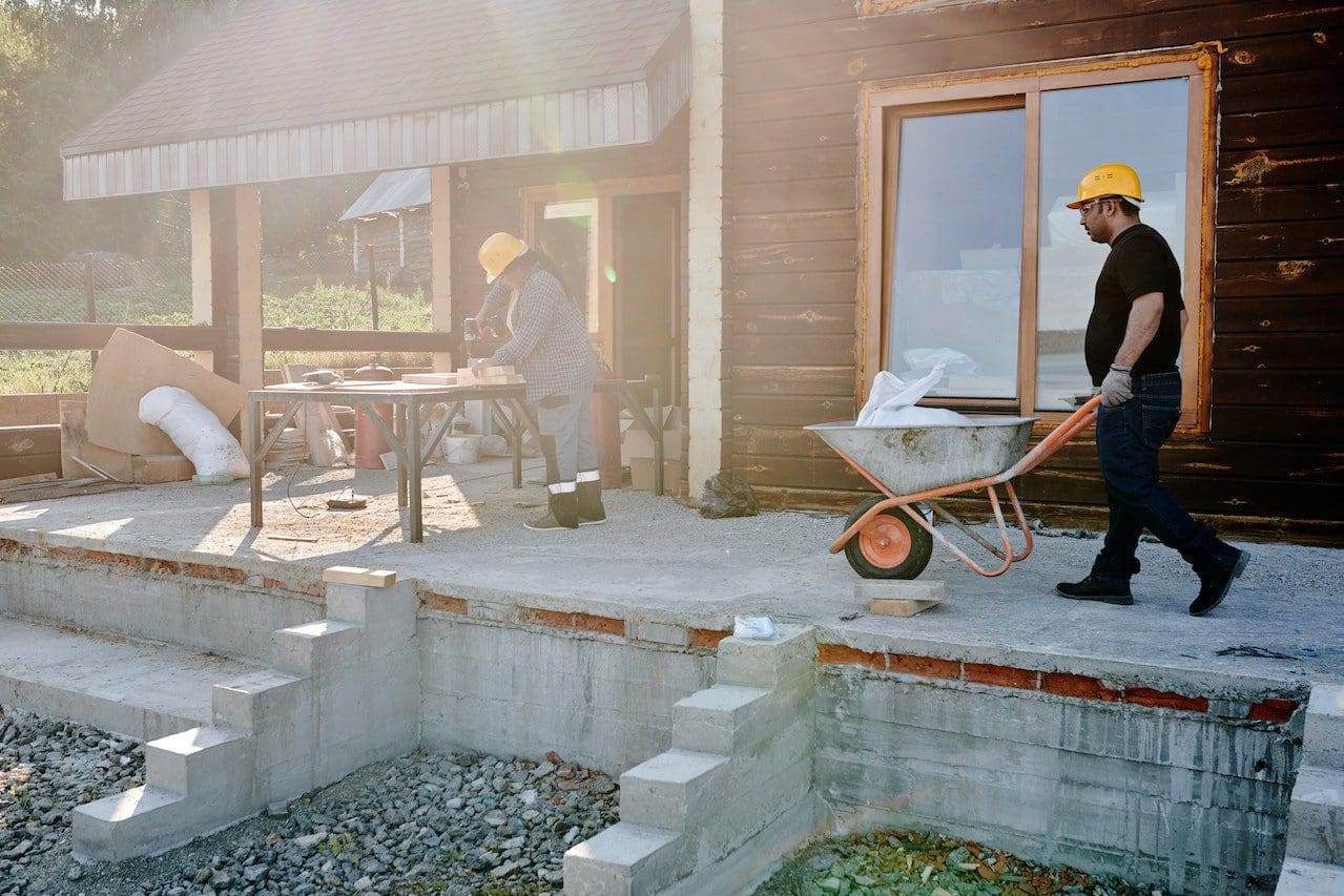 A man in yellow hard hat and black jacket using a wheelbarrow.